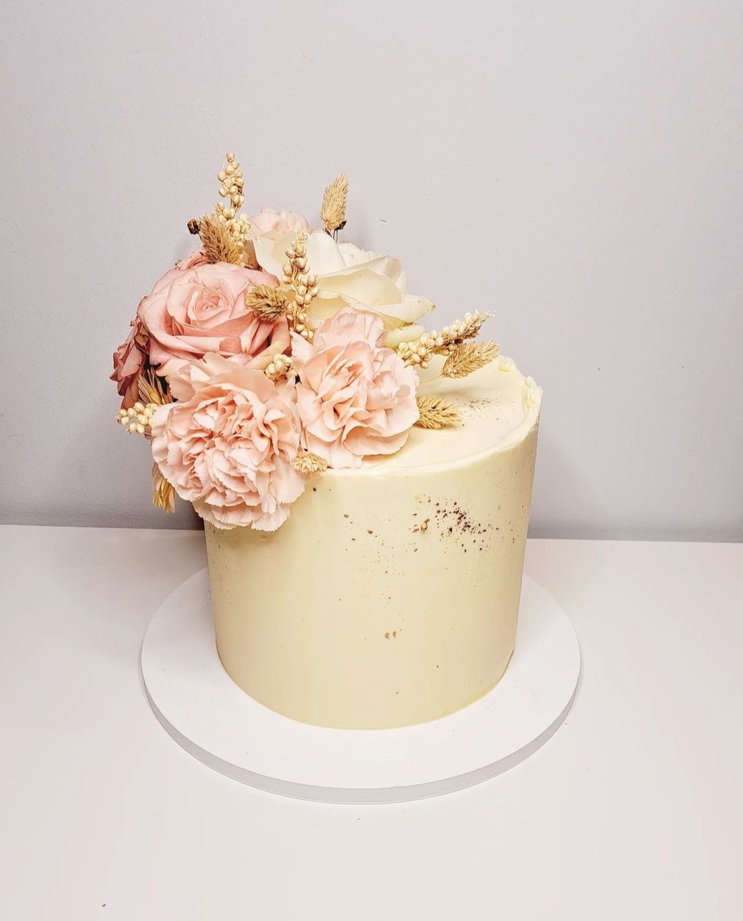 Tort z kremową polewą, ozdobiony kompozycją z różowych i białych kwiatów oraz suszonych traw, umieszczony na białym podeście.
