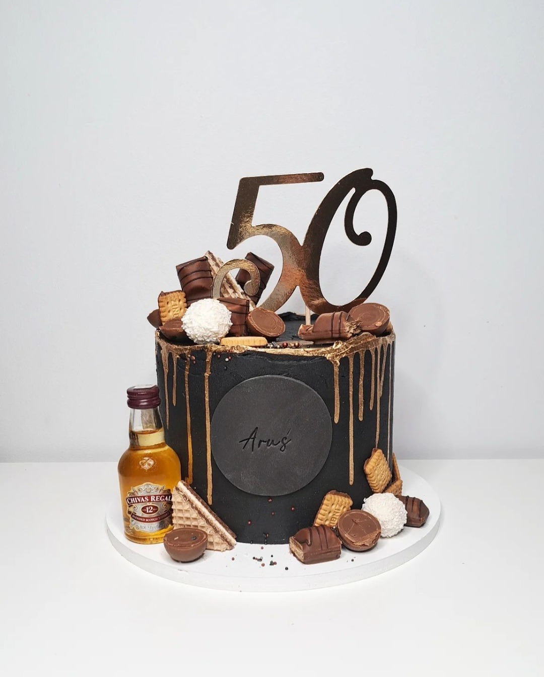 Czarny tort urodzinowy z złotymi akcentami, dekorowany czekoladkami, waflem, kokosowymi kulkami i miniaturową butelką whisky Chivas Regal, z topperem '50' i napisem 'Aruś'.