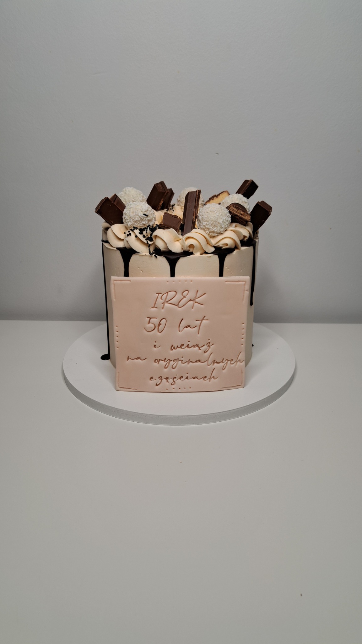 Tort urodzinowy z dekoracjami z czekolady, kulek kokosowych i tabliczką z napisem 'IREK 50 lat i wciąż na oryginalnych częściach' na białym tle.