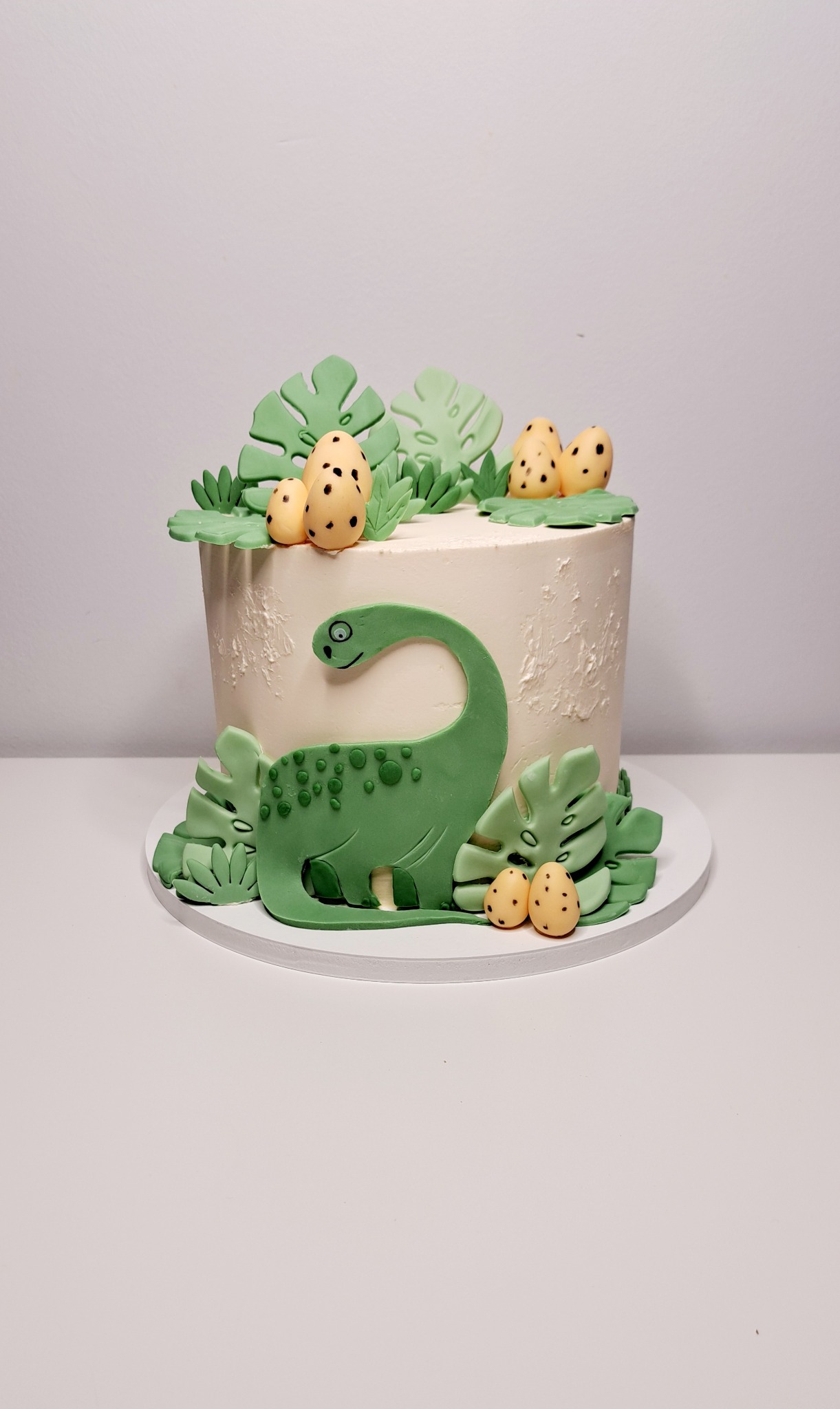 Tort z motywem dinozaura, dekorowany zielonymi liśćmi i jajkami z brązowymi plamkami, na białym tle.