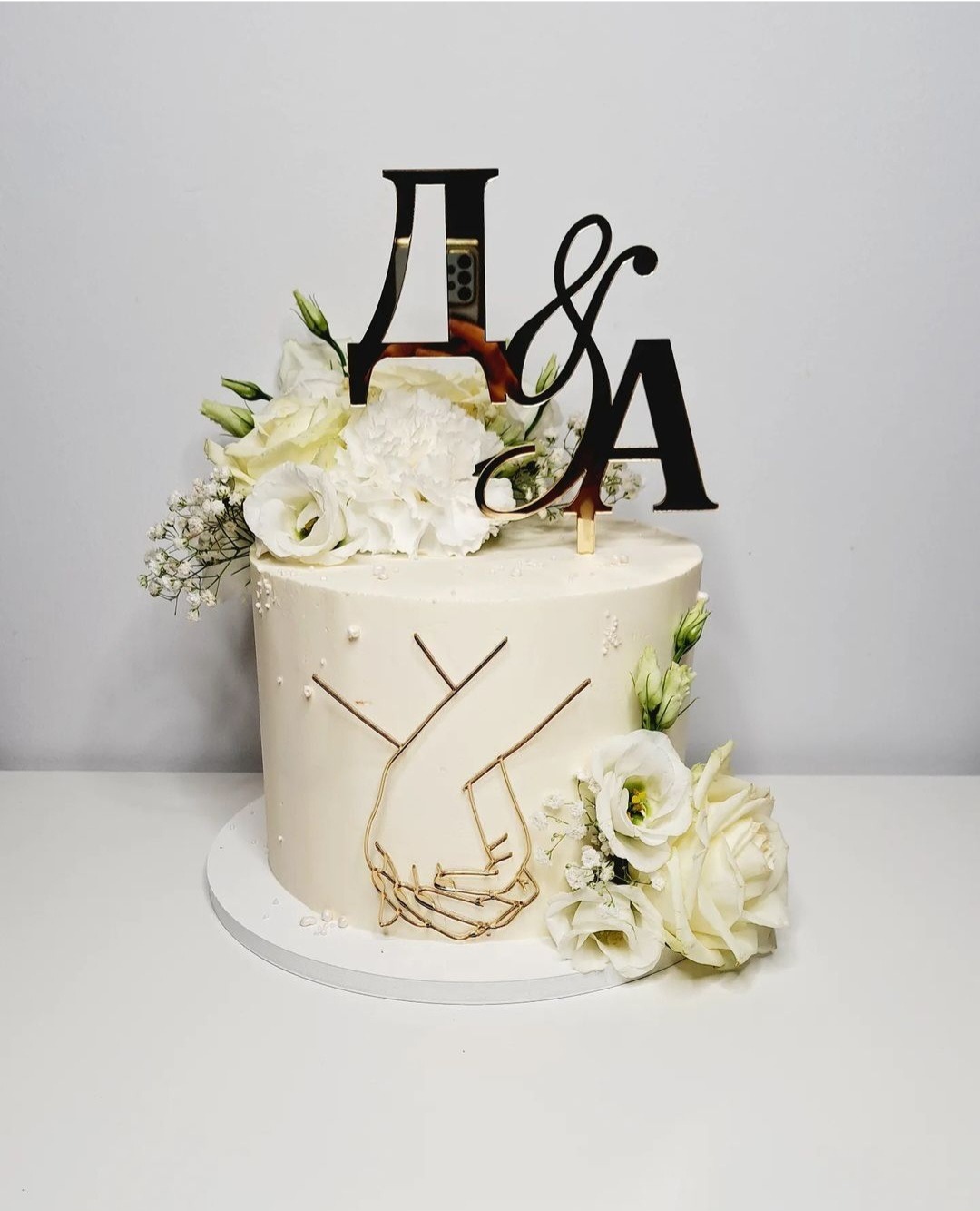 Elegancki tort weselny z białym kremem, dekoracją z białych kwiatów, topperem z inicjałami i motywem splecionych dłoni.