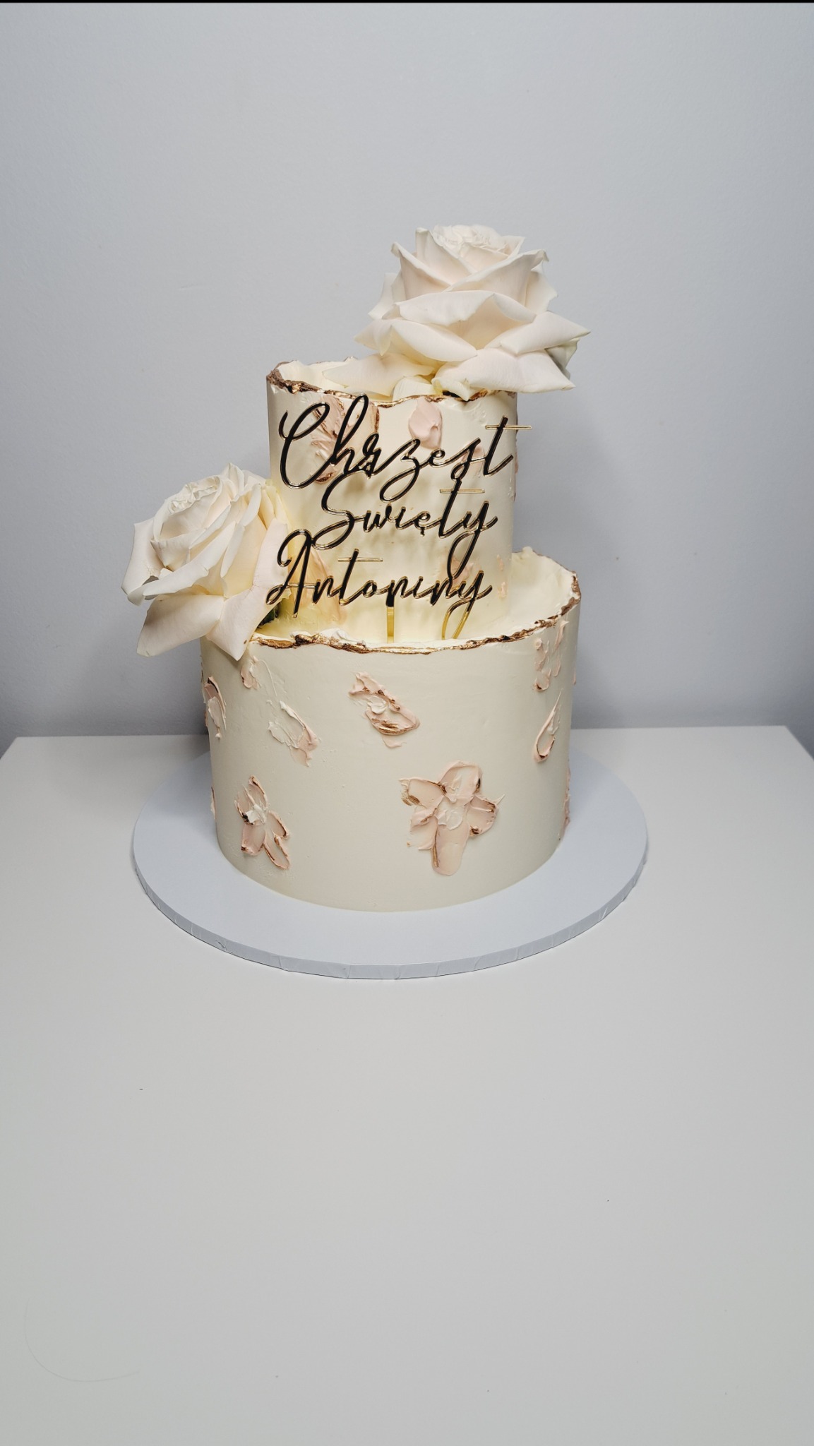 Dwupiętrowy tort w odcieniach bieli i różu, zdobiony kremowymi kwiatami i topperem z napisem 'Chrzest Święty Antoni', zwieńczony dwiema białymi różami.