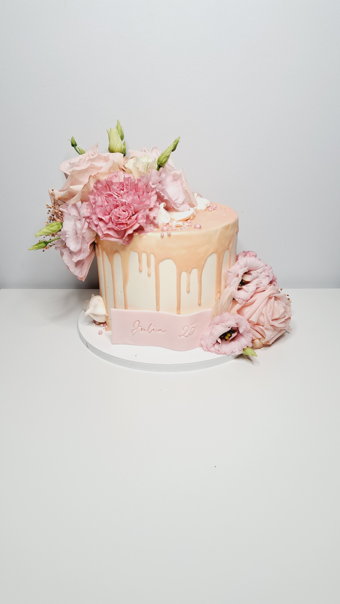 Tort urodzinowy w odcieniach różu, udekorowany świeżymi kwiatami i polewą, z napisem 'Julia 25' na dekoracyjnej wstążce.
