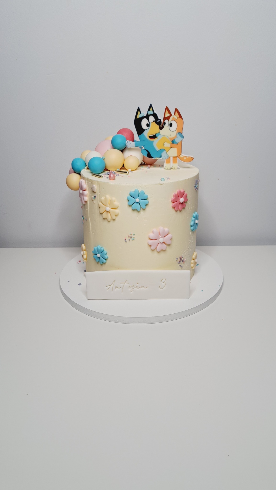 Tort urodzinowy z kremem, ozdobiony kolorowymi kwiatkami, pastelowymi kulkami i figurkami postaci z bajki Bluey, z napisem 'Antosia 3' na białej tabliczce.