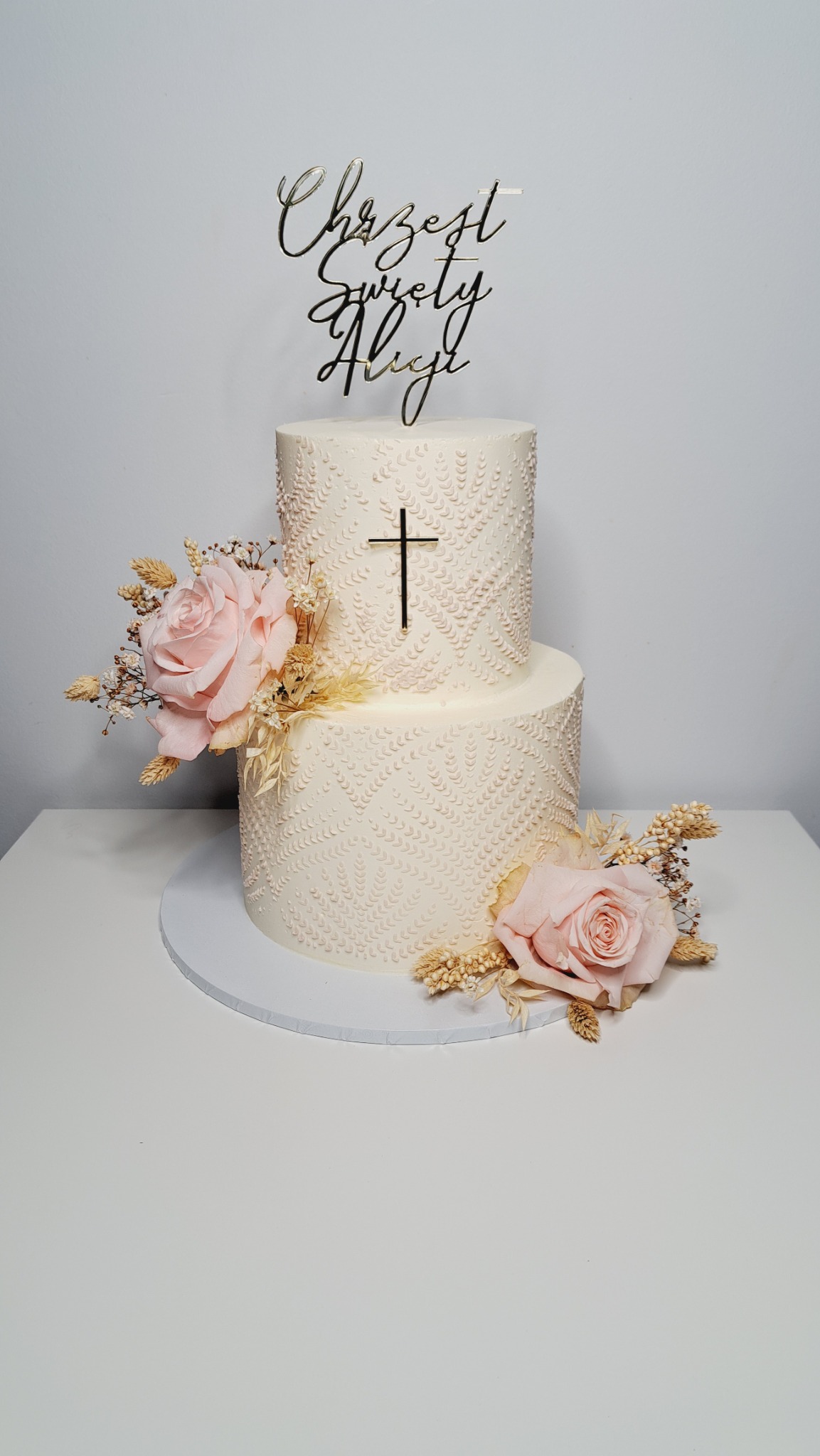 Dwupoziomowy tort w kremowym odcieniu, zdobiony wytłaczanym wzorem i krzyżem, z dekoracjami z różowych róż i suszonych traw, zwieńczony napisem 'Chrzest Święty Alicji' w złotym kolorze.