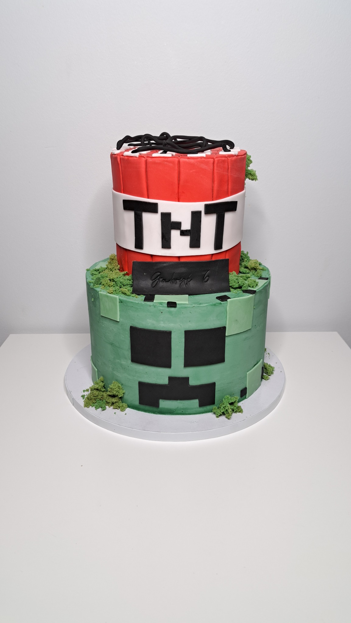 Dwupiętrowy tort w stylu Minecraft: górna warstwa przypomina dynamit TNT z czarnymi lontami, a dolna przedstawia zieloną twarz Creepera z czarnymi kwadratami. Dodatkowo tort udekorowany jest...