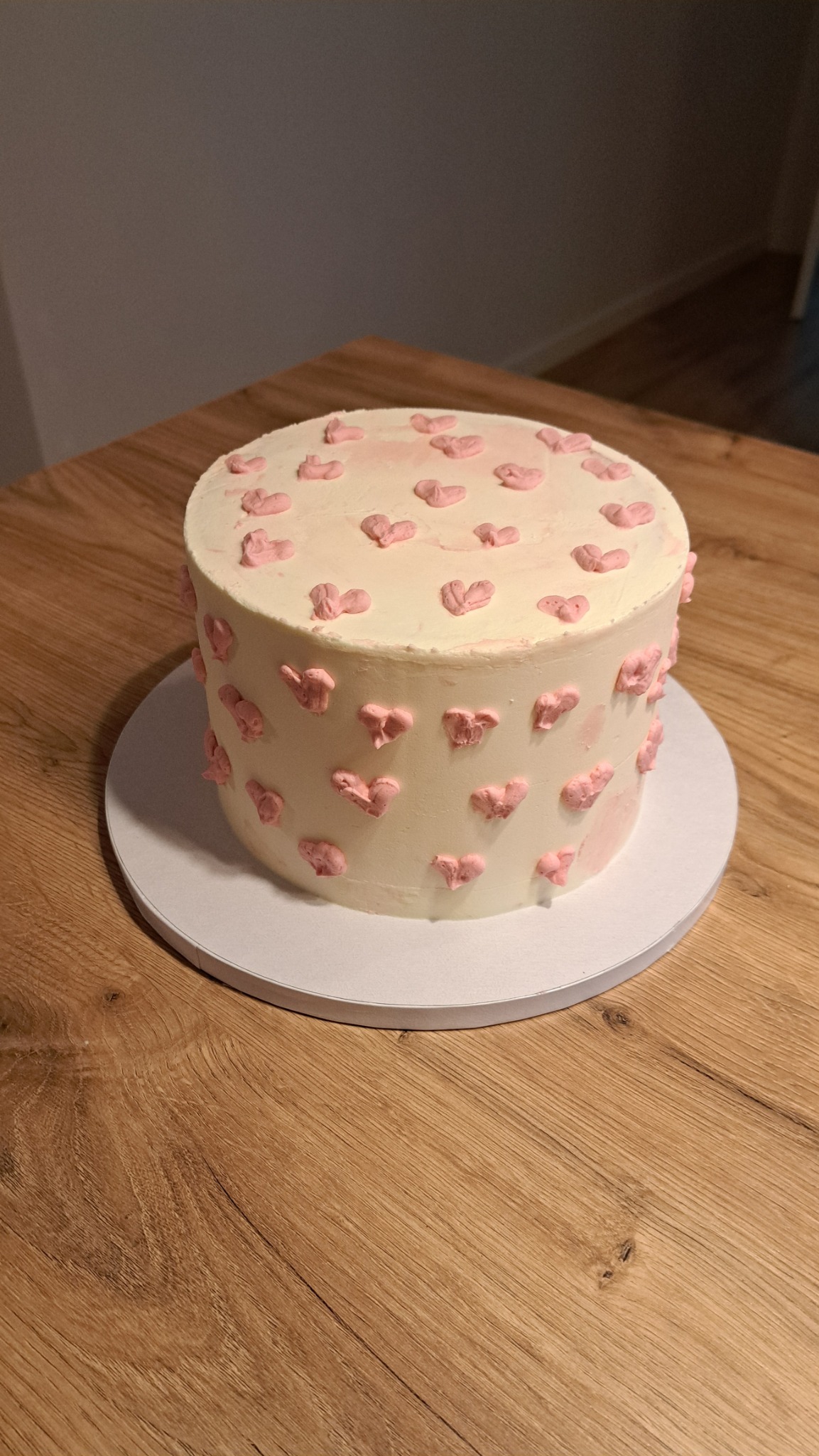 Tort z kremem w kolorze ecru, ozdobiony drobnymi, różowymi serduszkami, umieszczony na białym podkładzie na drewnianym stole.