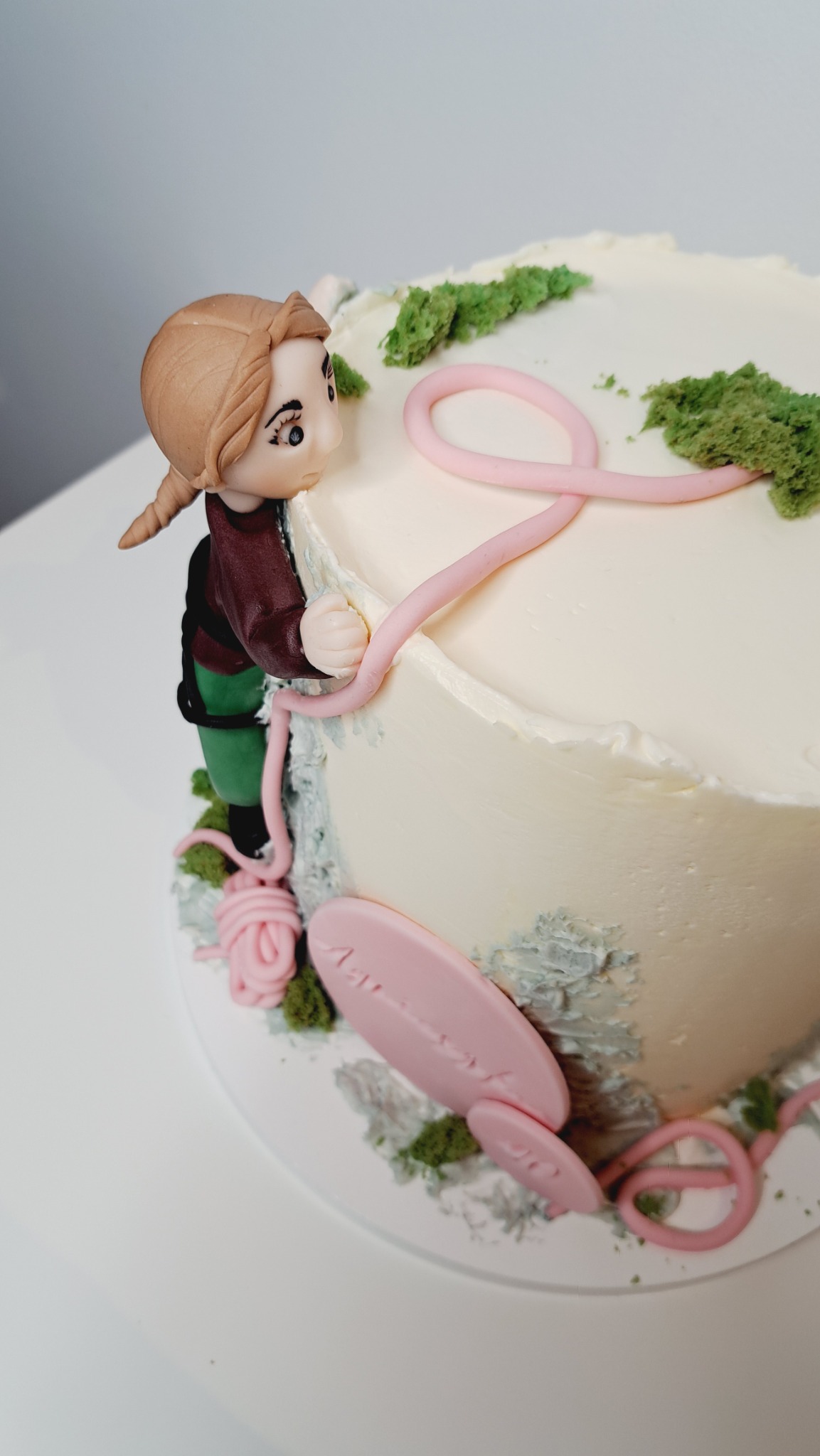 Tort zdobiony figurką dziewczynki z warkoczem, elementami przypominającymi włóczkę i zielonymi akcentami imitującymi roślinność, na białym tle.