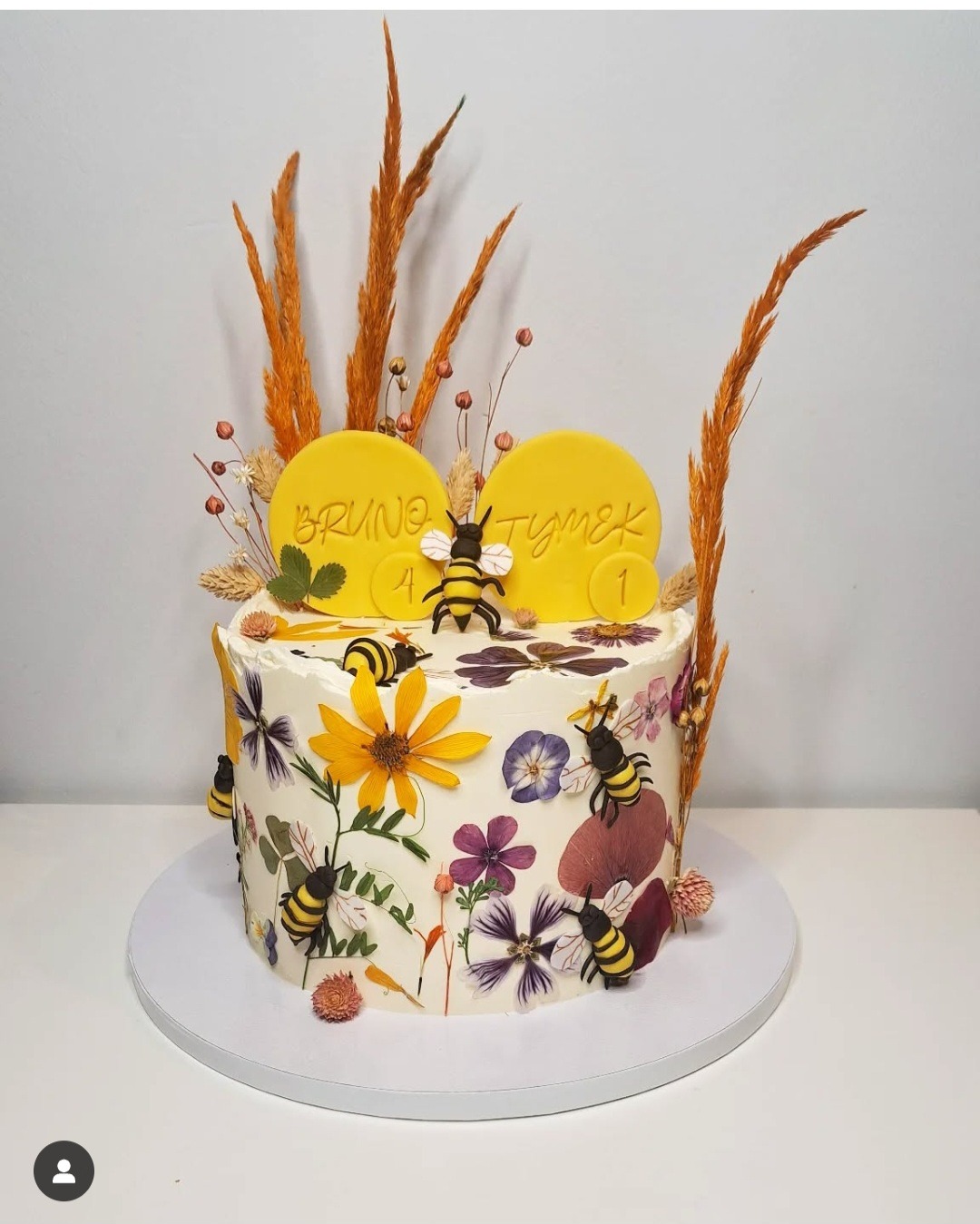 Tort urodzinowy ozdobiony suszonymi kwiatami, figurkami pszczół i żółtymi krążkami z imionami Bruno i Tymek, całość na białym tle.