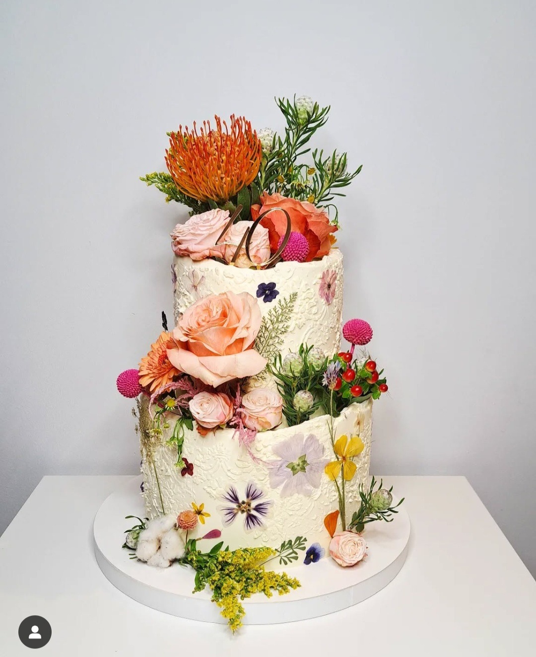 Dwupiętrowy tort weselny z kremem o fakturze koronki, udekorowany świeżymi kwiatami w odcieniach pomarańczy, różu i fioletu oraz złotym napisem '40'.