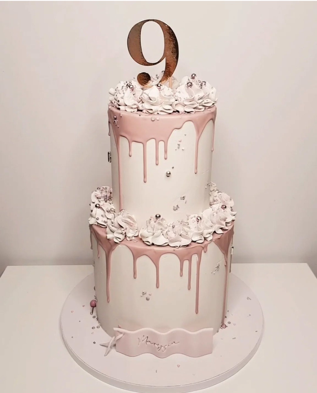 Dwupiętrowy tort urodzinowy z białą polewą, różowym lukrem spływającym po bokach, srebrnymi koralikami i topperem w kształcie cyfry 9 w kolorze różowego złota, dodatkowo zdobiony kremowymi...