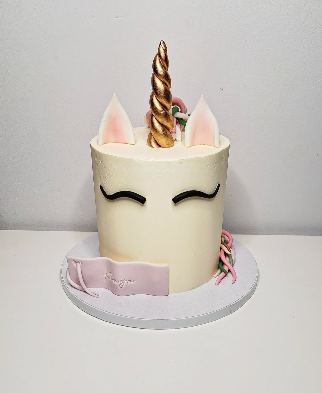 Tort jednorożec z kremowym lukrem, złotym rogiem i uszami z różowym detalem, z imieniem 'Kinga' na różowej wstążce.