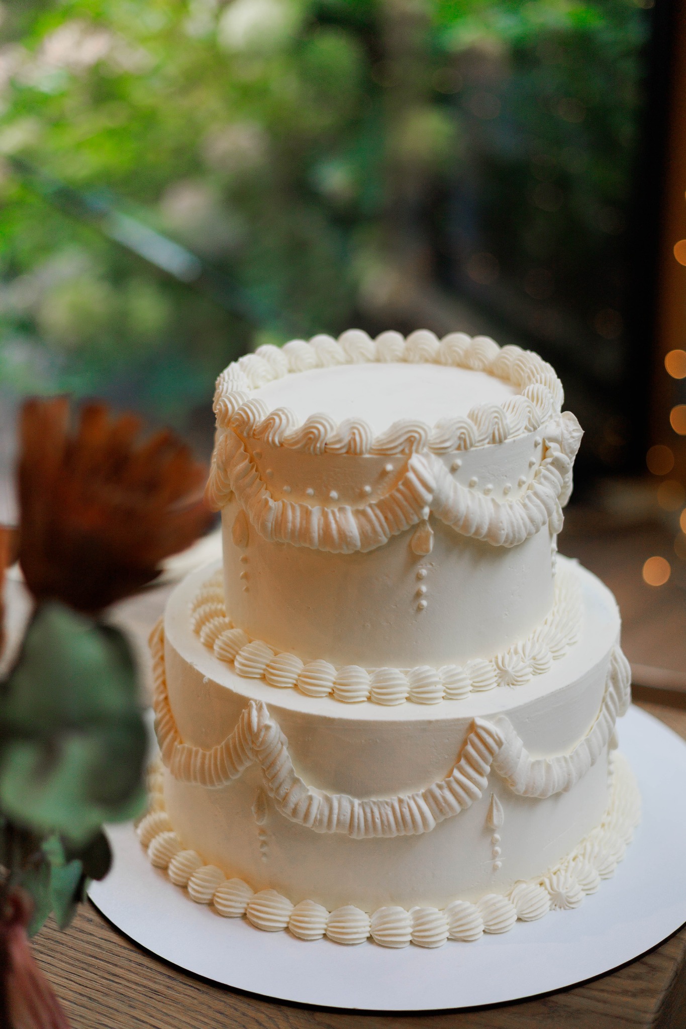 Dwupoziomowy tort weselny z kremową dekoracją w stylu vintage, na drewnianym stole, z rozmytym tłem zieleni i światełek, ukazujący precyzję wykonania.