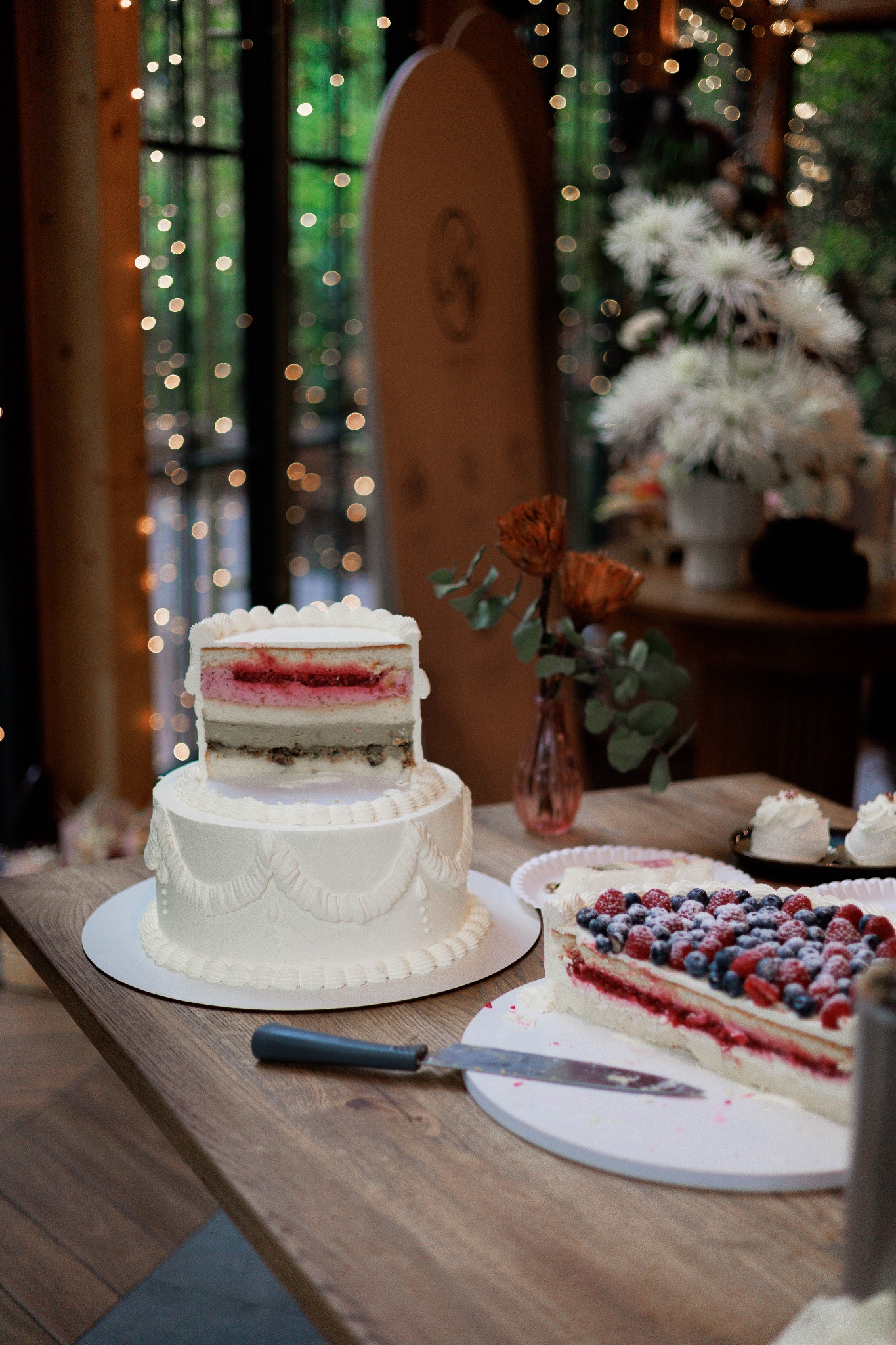 Dwupoziomowy tort weselny z przekrojem i kawałek tortu z owocami na drewnianym stole, w tle dekoracje świetlne i kwiaty.