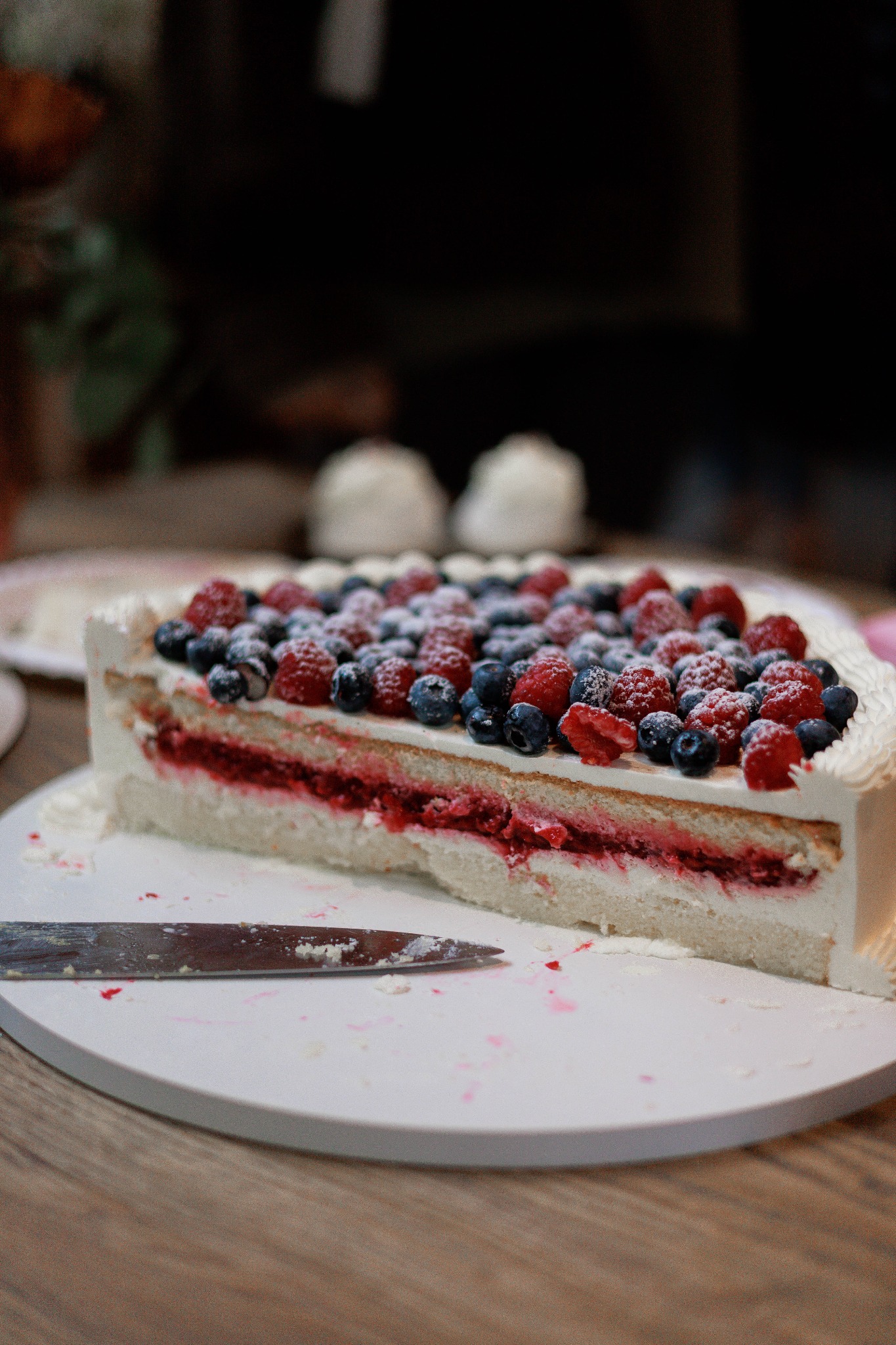Kawałek tortu z kremem, malinami i borówkami, posypany cukrem pudrem, leżący na białym talerzu obok noża. Widoczne ślady po krojeniu.