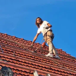 Pracownik z zabezpieczeniem na dachu w Gorzowie Wielkopolskim, montujący metalowe profile pod instalację paneli słonecznych na tle błękitnego nieba, widok z dołu na drabinę opartą o dach.