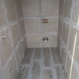 Wnętrze łazienki w trakcie prac wykończeniowych; widoczne płyty gipsowo-kartonowe na ścianach z zaszpachlowanymi łączeniami, przygotowane do dalszych prac, oraz częściowo ułożona podłoga z paneli...