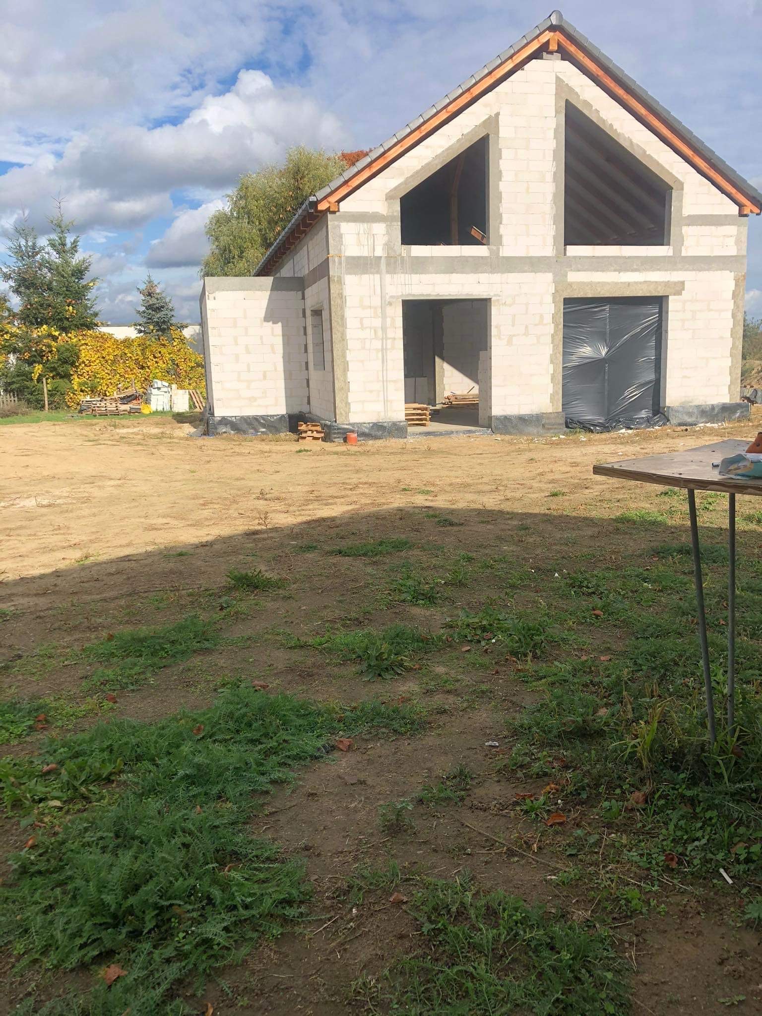 Surowy stan domu jednorodzinnego z pustaków, z widoczną konstrukcją dachu i folią zabezpieczającą otwór okienny, na działce z piaskiem i trawą.