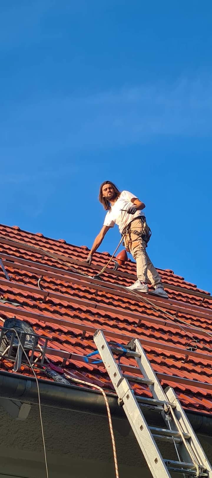 Pracownik z zabezpieczeniem na dachu w Gorzowie Wielkopolskim, montujący metalowe profile pod instalację paneli słonecznych na tle błękitnego nieba, widok z dołu na drabinę opartą o dach.