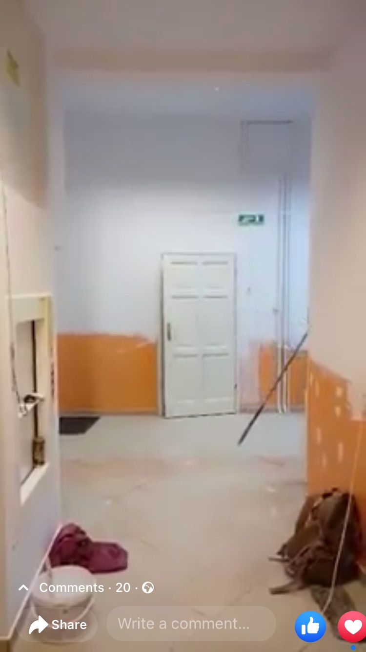 Korytarz biurowy w trakcie malowania, zabezpieczone dolne partie ścian pomarańczową folią, białe ściany i drzwi, na podłodze wiadro z farbą i torba narzędziowa.