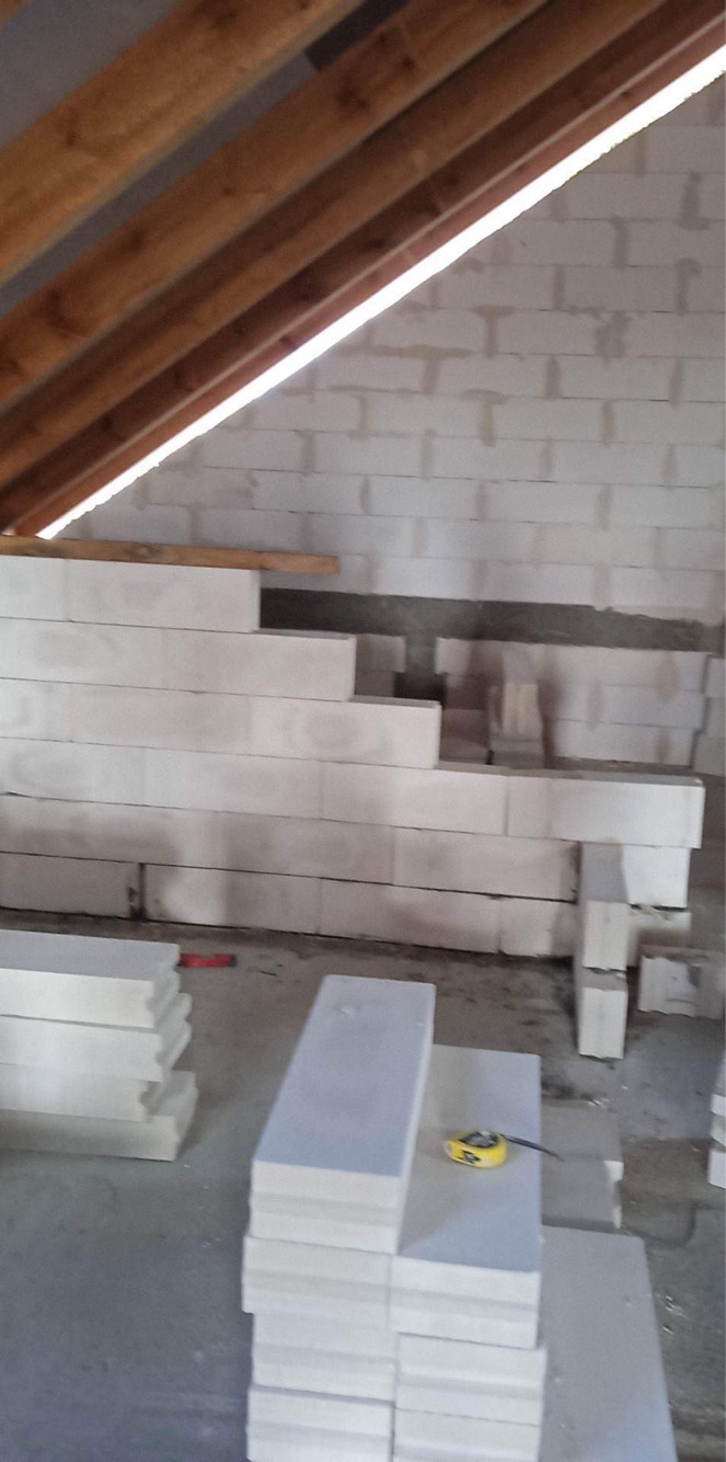 Poddasze w trakcie ocieplania, widoczne drewniane belki stropowe, ściany z białego bloczka i ułożone warstwami płyty izolacyjne z widoczną miarą zwiniętą na jednej z nich.