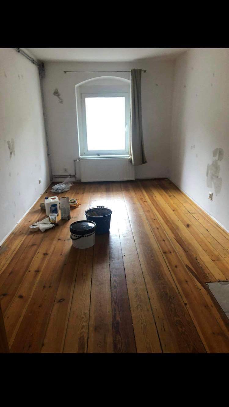 Pokój w trakcie przygotowań do prac wykończeniowych: drewniana podłoga po cyklinowaniu, ściany z widocznymi śladami po naprawach, wiaderka z farbą i akcesoria malarskie przy ścianie.