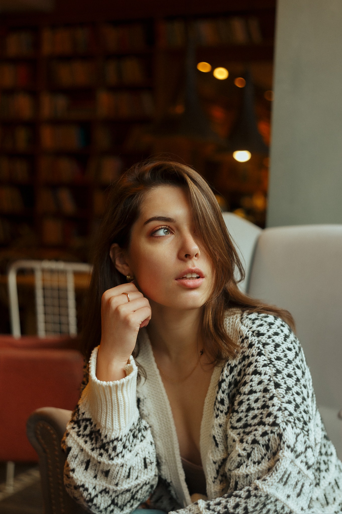 Portret kobiety z długimi brązowymi włosami, w swetrze z czarno-białym wzorem, siedzącej w fotelu na tle regału z książkami. Uwagę przykuwa złoty pierścionek na jej palcu i zamyślone spojrzenie w bok.