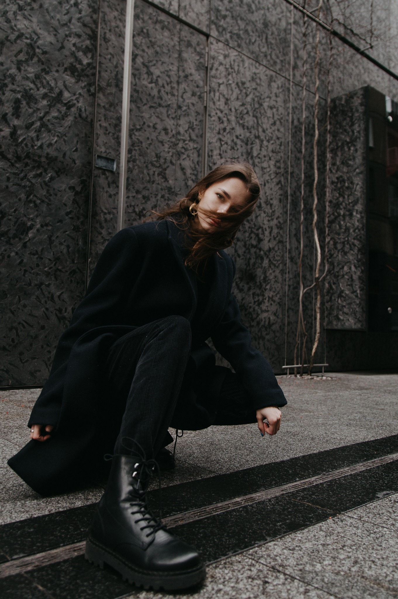 Kobieta w czarnym płaszczu i butach sznurowanych, pozująca na tle grafitowej ściany z fakturą, włosy rozwiane wiatrem, złote kolczyki koła.