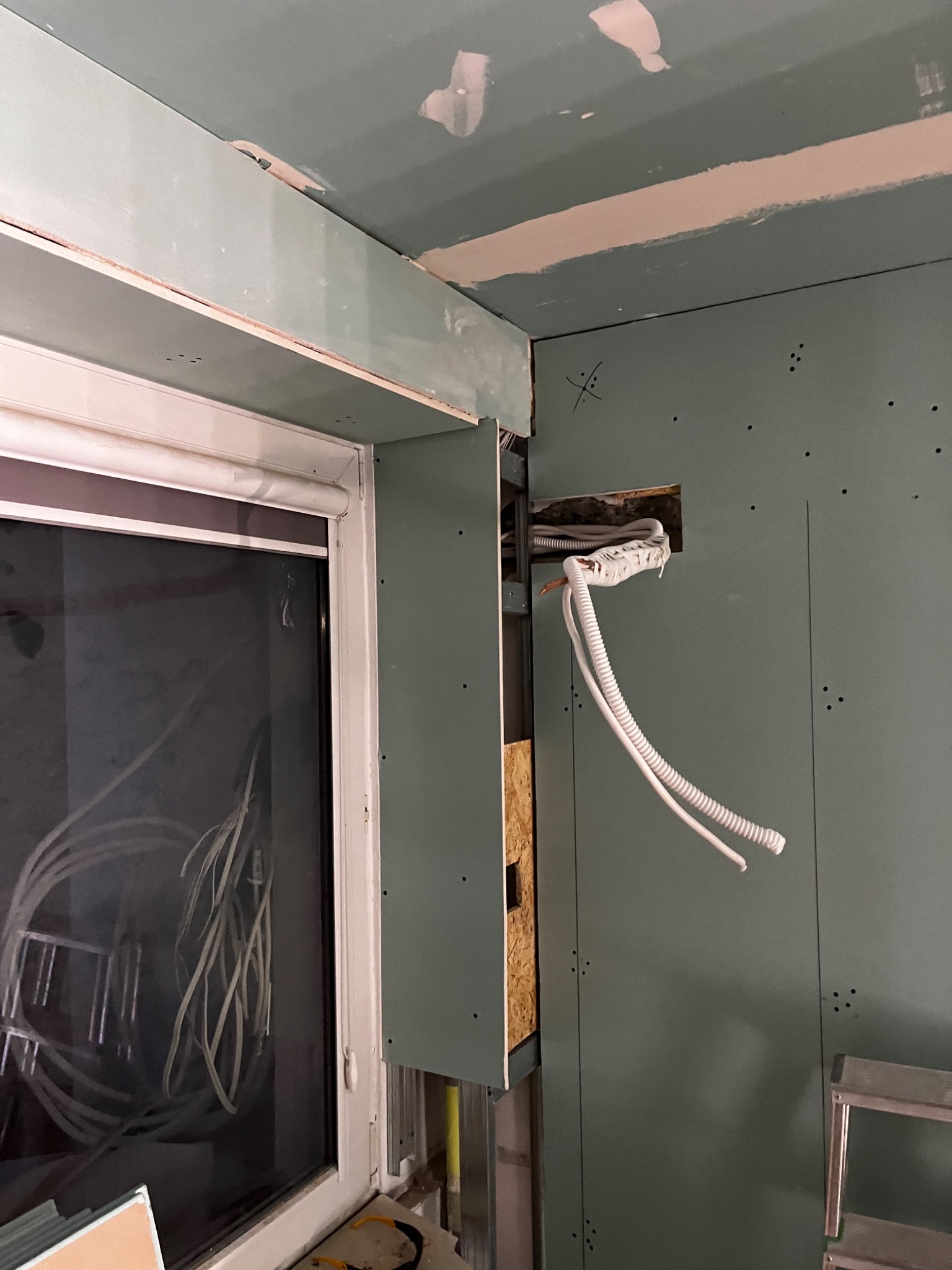 Montaż płyt gipsowo-kartonowych w pomieszczeniu, widoczne przewody elektryczne wystające ze ściany, okno z roletą i częściowo zamontowane płyty na suficie.