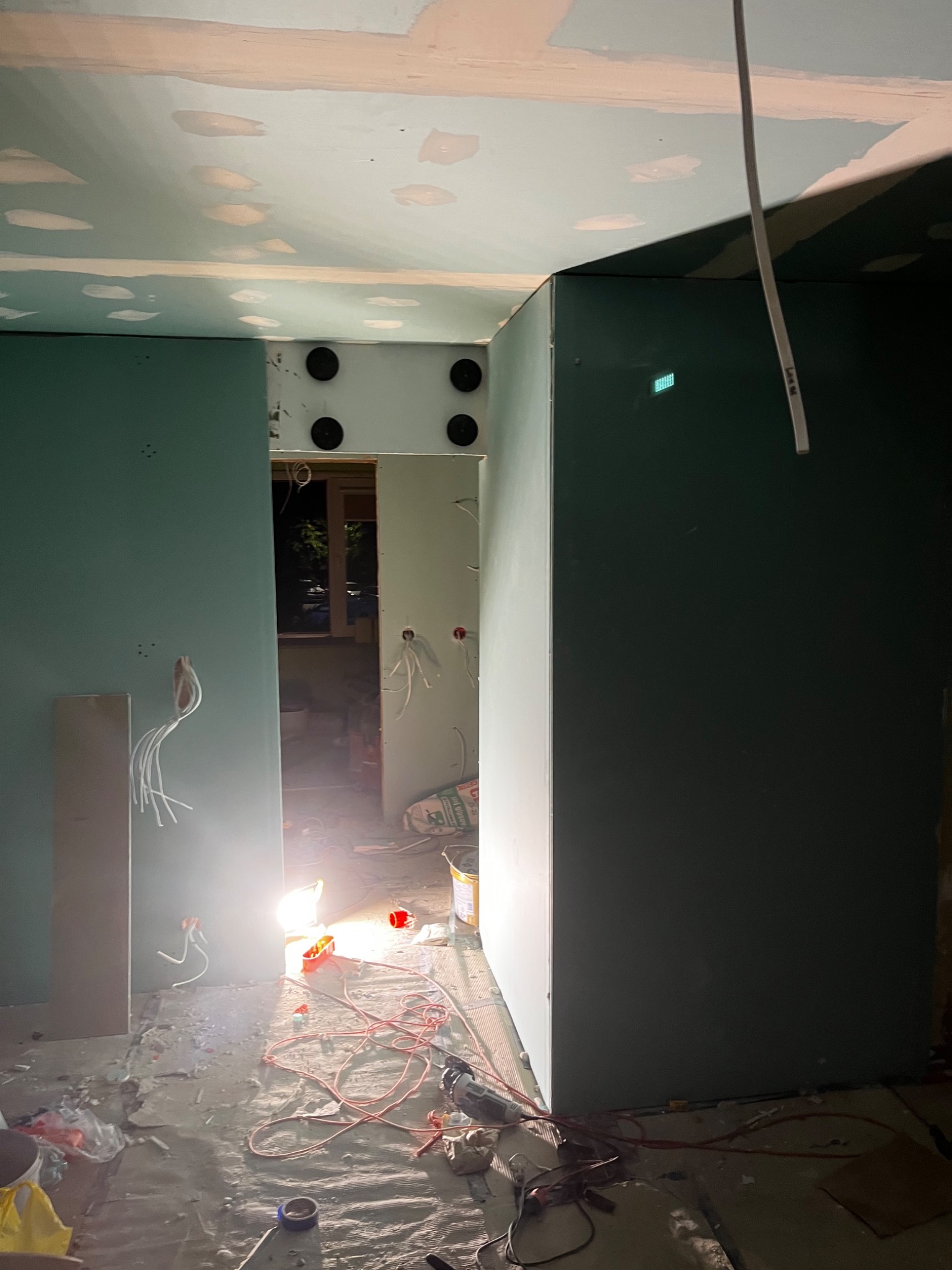 Wnętrze w trakcie montażu płyt gipsowo-kartonowych, widoczne przewody elektryczne, narzędzia i resztki materiałów na podłodze, sufit częściowo wykończony z widocznymi miejscami po szpachlowaniu.