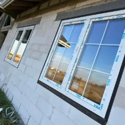 Nowe okna z białymi ramami, marki Veka, zamontowane w surowej ścianie z bloczków, widoczne z zewnątrz budynku w trakcie budowy.