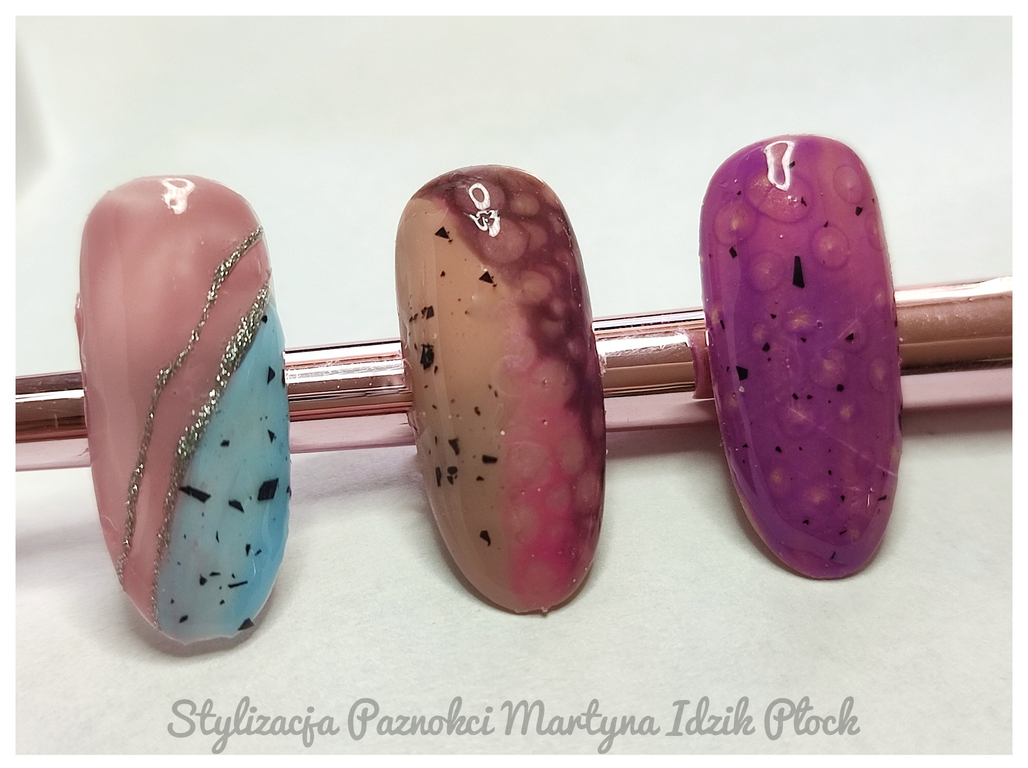 Trzy sztuczne paznokcie z różnymi stylizacjami: geometryczny wzór z brokatem, ombre z czarnymi drobinkami i fioletowy z efektem kropli.