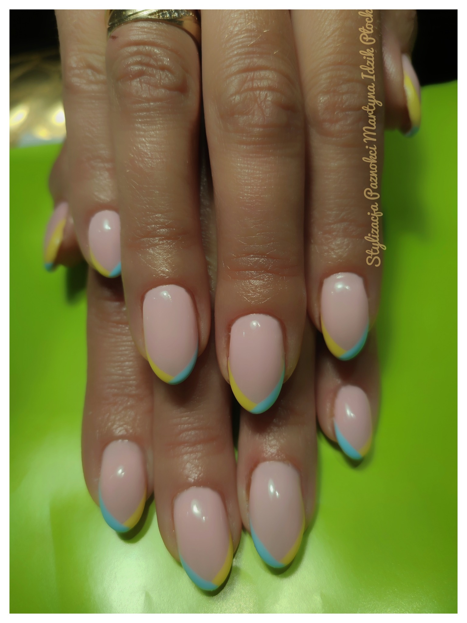 Dłonie z pastelowym różowym manicure, zdobione żółto-błękitnymi końcówkami na zielonym tle.