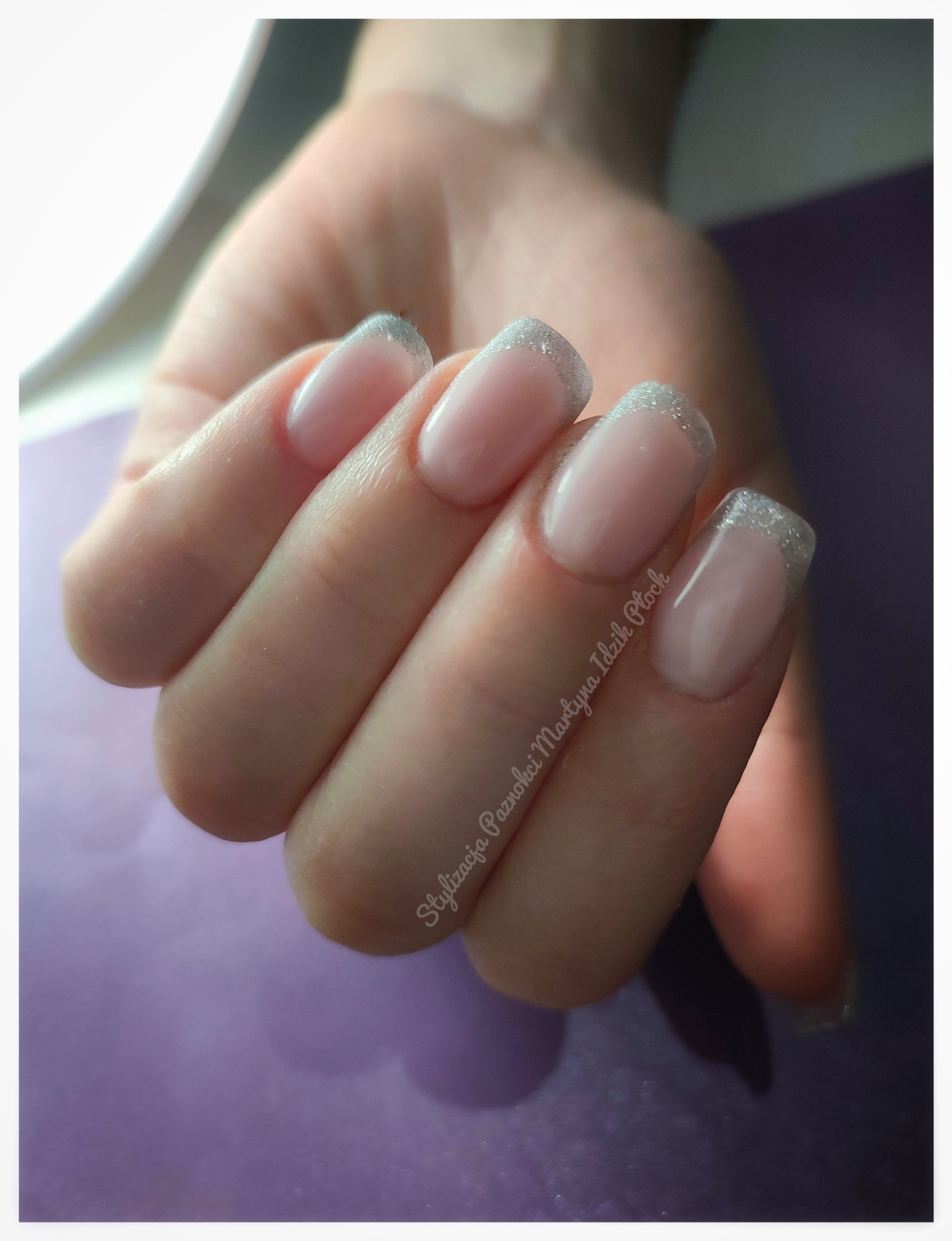 Dłoń z delikatnym, błyszczącym french manicure z użyciem srebrnego brokatu na końcówkach paznokci, na fioletowym tle, z widocznym napisem 'Stylizacja Paznokci Martyna Idzik Płock'.