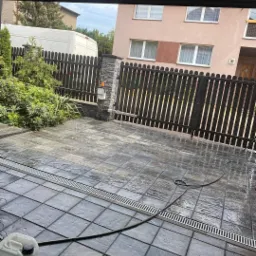 Ułożona szara kostka brukowa z odpływem liniowym i wężem ogrodowym, widoczny fragment ogrodzenia i budynek w tle.