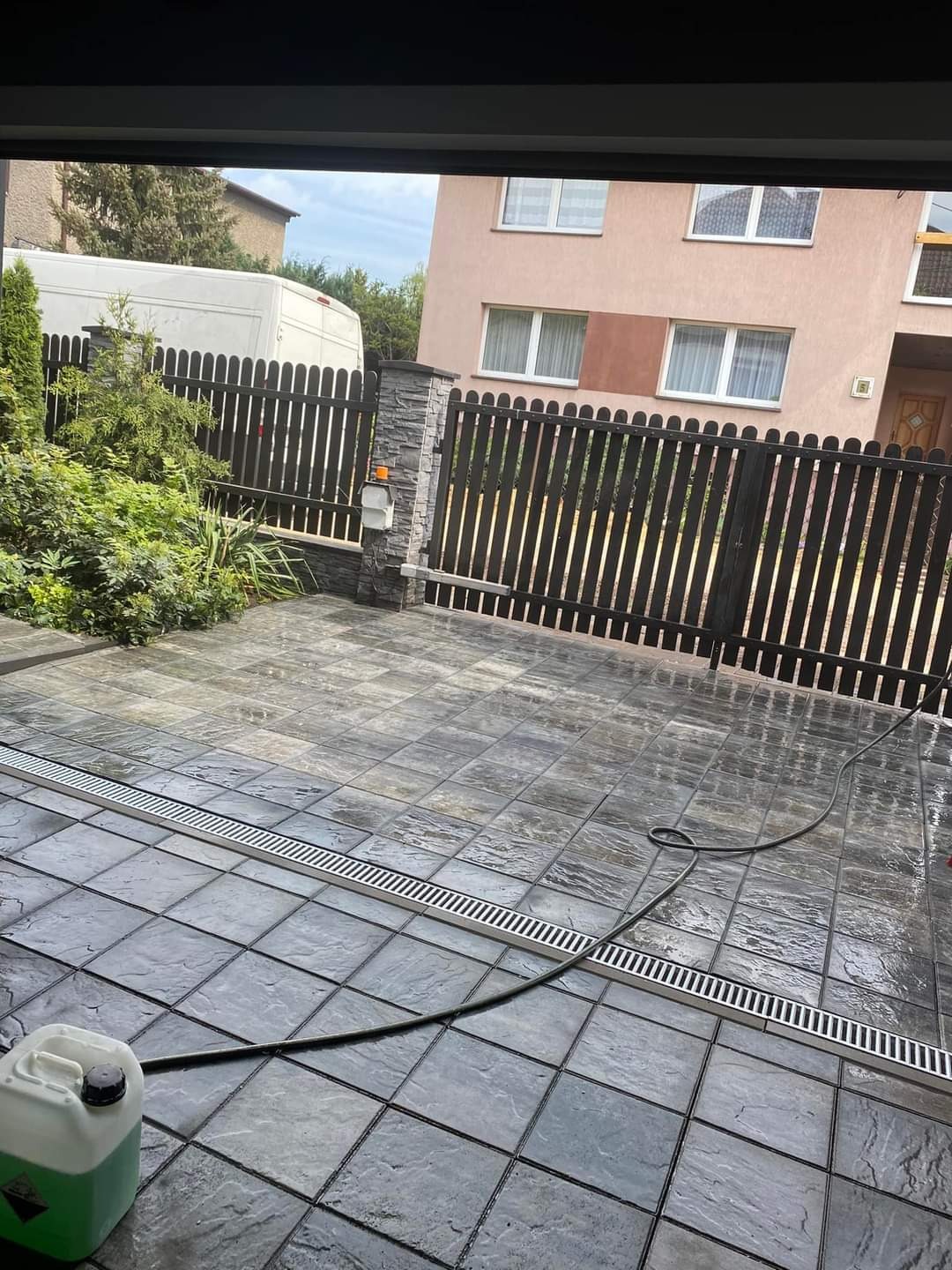 Ułożona szara kostka brukowa z odpływem liniowym i wężem ogrodowym, widoczny fragment ogrodzenia i budynek w tle.