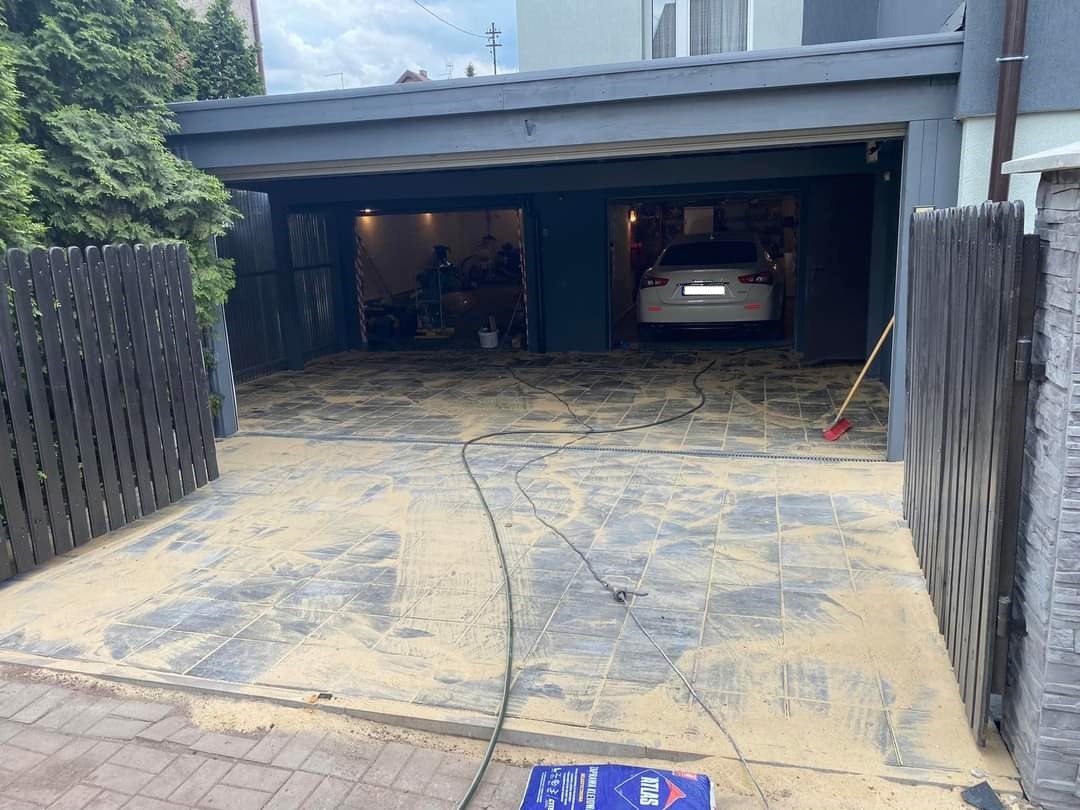Układanie kostki brukowej na podjeździe do garażu w Chorzowie, widoczny piasek kwarcowy do wypełniania szczelin, częściowo zasypana kostka, w tle zaparkowany samochód w garażu.