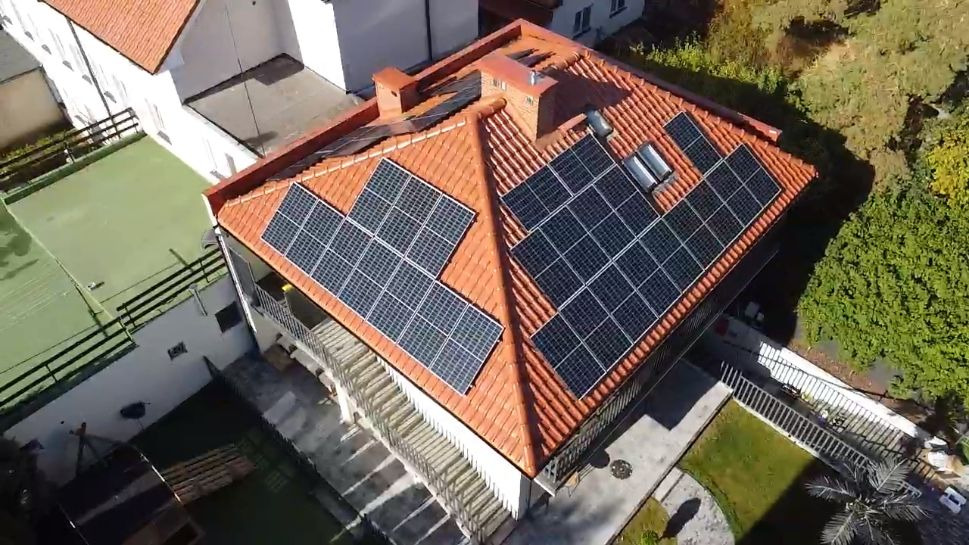 Widok z góry na dom z pomarańczową dachówką, na której zamontowano panele słoneczne w kilku sekcjach, otoczony zielenią.