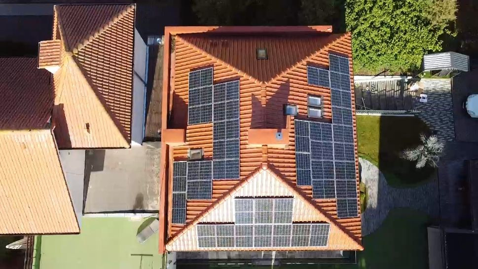 Widok z drona na dach domu z pomarańczowej dachówki, na którym zamontowane są panele fotowoltaiczne w różnych układach, otoczony zielenią i innymi budynkami.