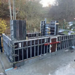 Zalany betonem szalunek fundamentów z widocznymi prętami zbrojeniowymi i elementami mocującymi, otoczony drzewami w jesiennej scenerii.