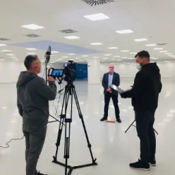 Ekipa filmowa w maskach nagrywa wywiad z mężczyzną w garniturze w przestronnym, jasnym pomieszczeniu z równomiernym oświetleniem sufitowym.