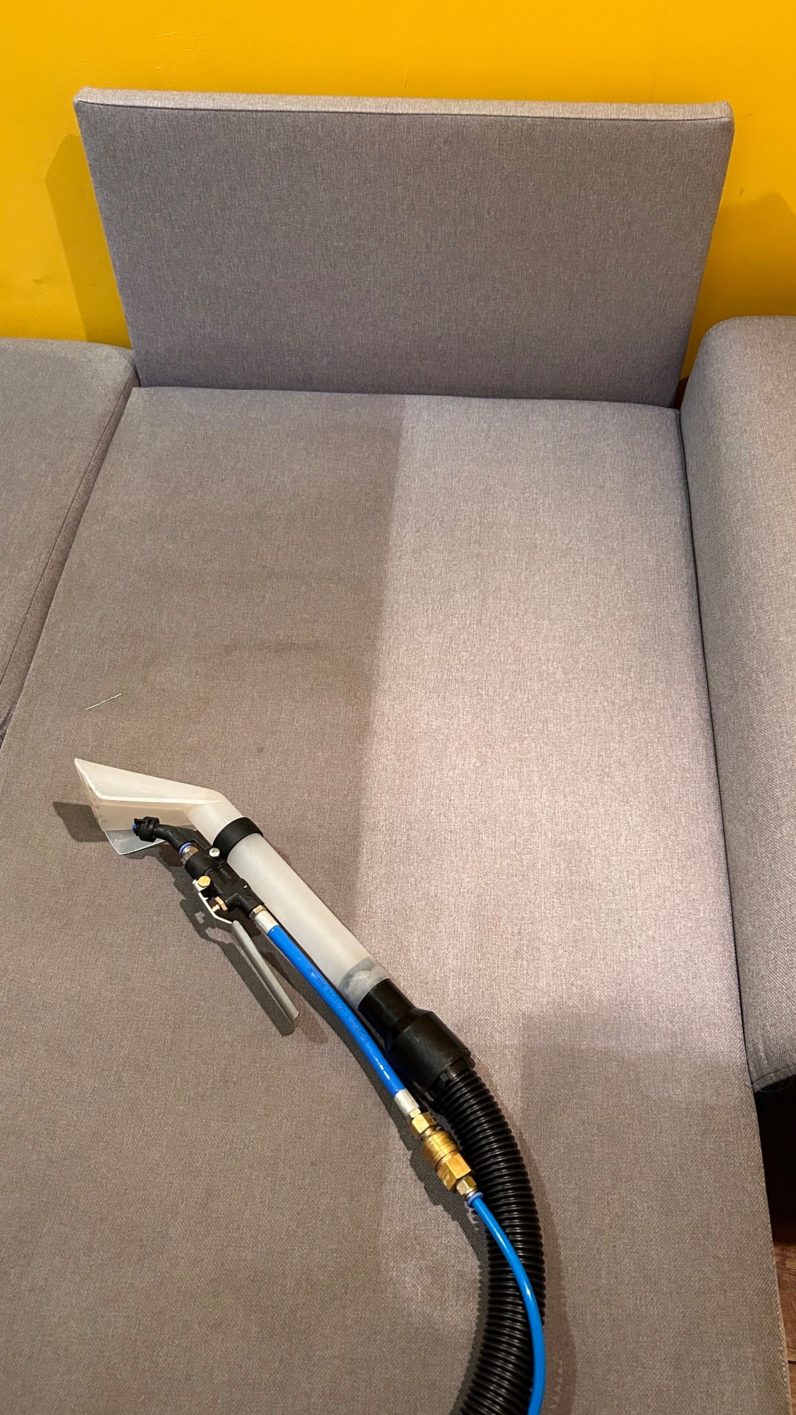 Fragment szarej sofy, widoczna różnica w odcieniu po czyszczeniu, z dyszą odkurzacza piorącego na powierzchni, żółta ściana w tle.