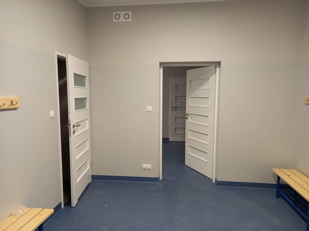Szara szatnia z niebieską podłogą, widoczne dwie otwarte białe drzwi z przeszkleniami i drewniane ławki pod ścianami.