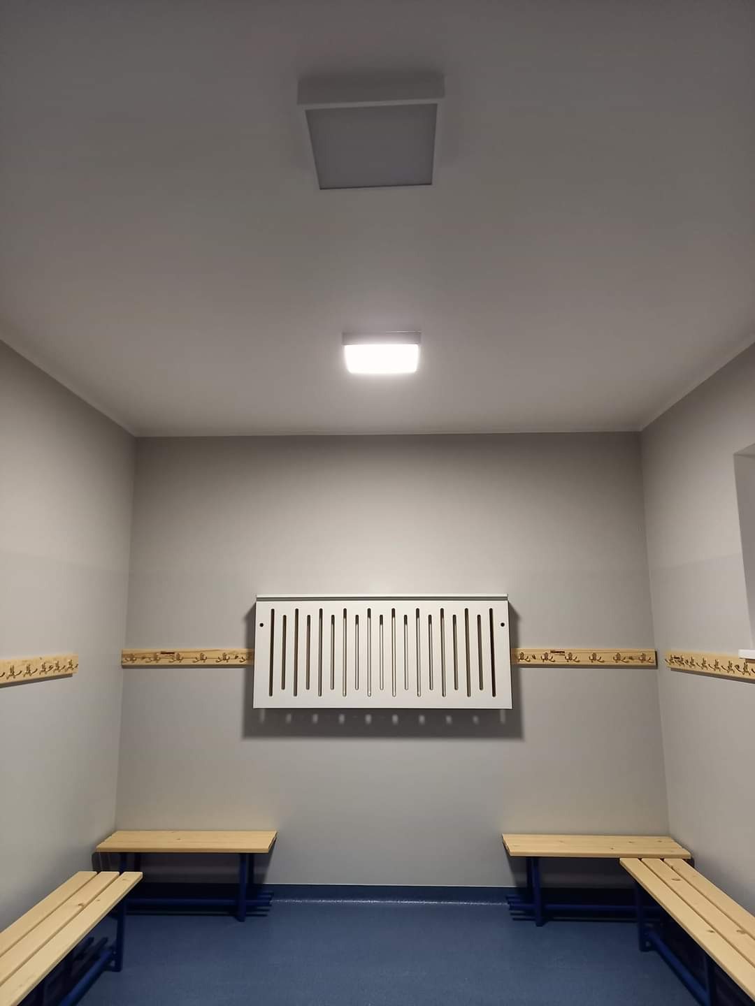 Wnętrze szatni z niebieską podłogą, drewnianymi ławkami i wieszakami na ścianach, oświetlone dwoma kwadratowymi lampami sufitowymi.