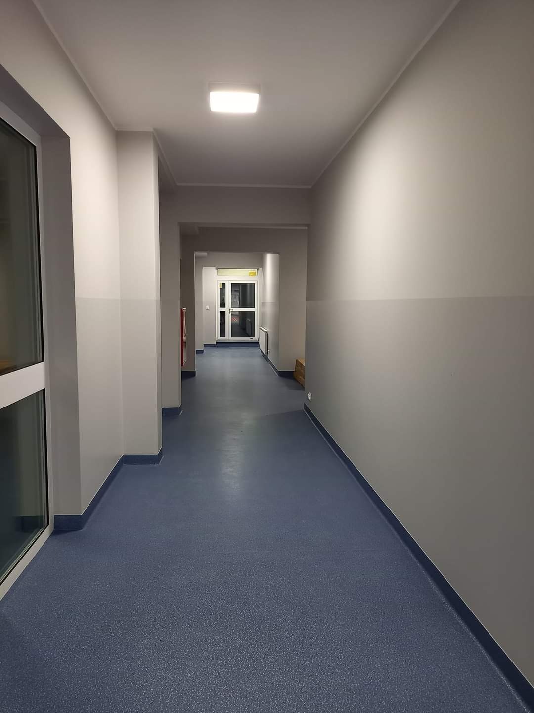 Długi korytarz z szarymi ścianami z ciemniejszym pasem na dole i niebieską podłogą, oświetlony przez lampę sufitową i światło z końca korytarza.
