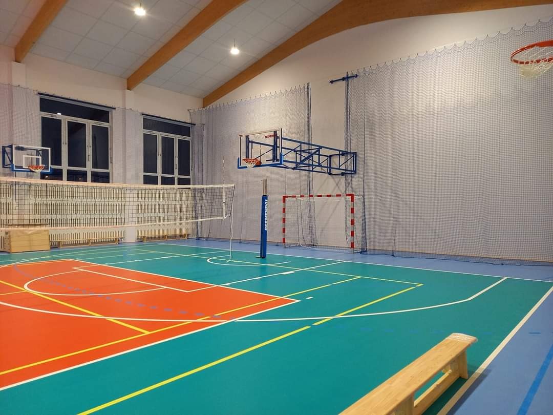 Wnętrze hali sportowej z parkietem w odcieniach niebieskiego i pomarańczowego, widoczne kosze do koszykówki, siatka do siatkówki oraz bramka do piłki ręcznej, oświetlenie sufitowe i drewniane belki...