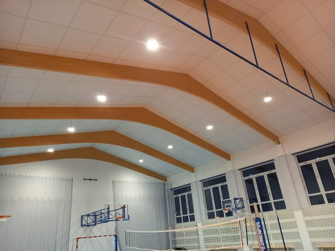 Wnętrze hali sportowej z drewnianą konstrukcją nośną dachu, białym sufitem kasetonowym i oświetleniem punktowym, widoczne kosze do koszykówki i siatka do siatkówki.