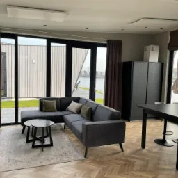 Nowoczesny salon z szarym narożnikiem, dwiema okrągłymi kawowymi stolikami i parkietem w jodełkę, widok na jezioro i domki letniskowe przez duże przesuwne okna.