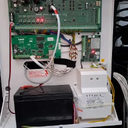 Otwarta obudowa centrali alarmowej z widocznymi modułami elektronicznymi, akumulatorem i transformatorem, okablowanie wewnątrz obudowy, widoczne diody LED sygnalizujące pracę urządzenia.