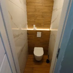 Wąska toaleta z podświetleniem LED między marmurkowymi i drewnopodobnymi płytkami, widok zza uchylonych drzwi.